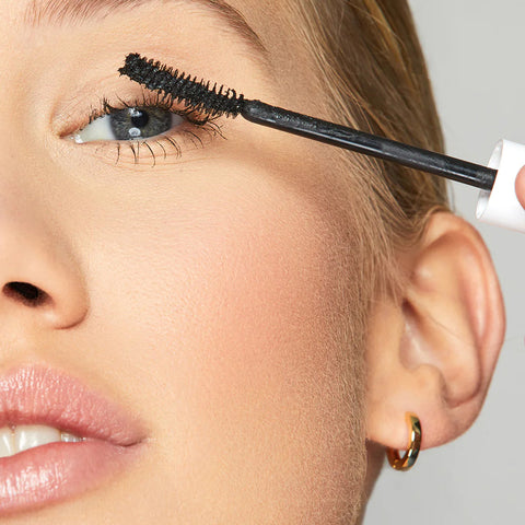 A person uses Twenty Twenty Clean Sweep Mascara in Pitch Black on their lashes, highlighting their eye. Its perfect for sensitive eyes and complements smooth skin and subtle makeup. The person also sports a small hoop earring and has slightly parted lips.