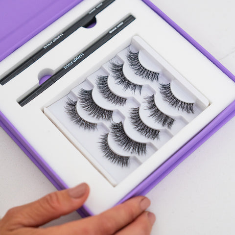 A hand holds a purple Merakim Eyes on Beauty Lashes box, showcasing five faux mink lash pairs and two eyeliner pencils, all elegantly arranged in white packaging for a flawless presentation.