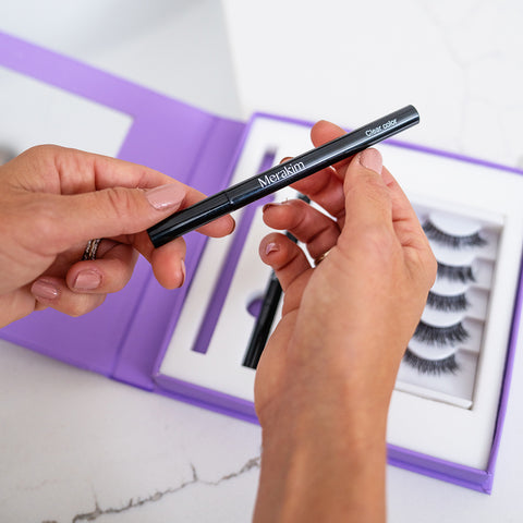 A person holds a black Merakim eyeliner pen labeled Clear Color. In the background, an open purple box reveals the Merakim Eyes on Beauty Lashes, featuring faux mink lashes on a white tray.