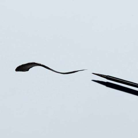 The Twenty Twenty Double Duty Precision Eyeliner in Pitch Black, lying against a light gray background, creates a swooping black line with a thick start and tapered end. Its ophthalmologist-tested for eye safety while beautifully accentuating your eyes.