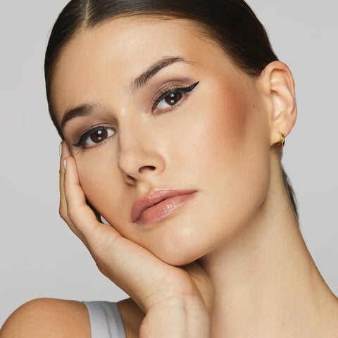 A woman poses with her hand on her face, showcasing sleek dark hair, glossy lips, light blush, and eye-safe winged eyeliner using Twenty Twentys Double Duty Precision Eyeliner in Pitch Black. Small gold hoops complement her look above a light gray tank top against a plain background.