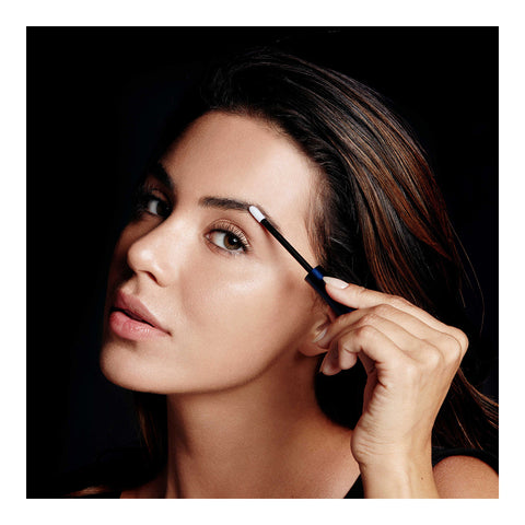 A person with long, dark hair is applying eyebrow makeup using a small brush and RevitaLashs RevitaBrow Advanced Eyebrow Conditioner Serum. The dark background highlights their face, with a focused look and glowing skin enhanced by the nourishing BioPeptin Complex® in the serum.