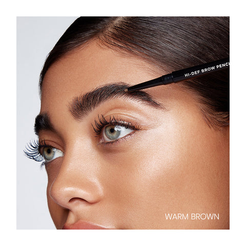 A close-up shows a person using the water-resistant, hypoallergenic RevitaLash Hi-Def Brow Pencil in Warm Brown to shape their eyebrow. The smooth skin and long lashes accentuate the well-defined, long-wear brow. WARM BROWN appears at the bottom right.