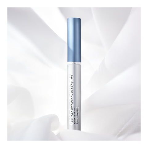 A cylindrical tube of RevitaLash Advanced Eyelash Conditioner and Enhancing Serum Sensitive features a blue cap, standing upright against a soft white fabric background. The product name is elegantly printed vertically, promoting enhanced eyelash health.