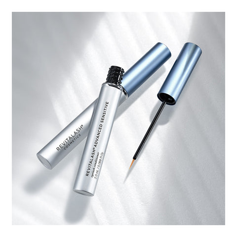 Two metallic blue tubes of RevitaLash Advanced Eyelash Conditioner and Enhancing Serum Sensitive are shown with their caps off. One tube has a fine-tipped applicator brush for precise application, ensuring optimal eyelash health. They rest on a smooth, light surface under bright lighting.