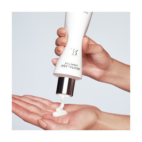 A person squeezes RevitaLash Cosmetics Thickening Conditioner Scalp Therapy Formula, a rich white cream, from a sleek white tube with a silver cap and subtle branding into their palm. The light gray background creates a clean, minimalistic look.