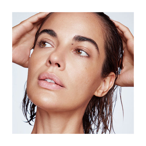 With wet hair enhanced by RevitaLashs Thickening Conditioner Scalp Therapy Formula, Hypoallergenic (250mL), and natural makeup, a person looks to the side touching their head. Their skin looks smooth and dewy against a plain white background.