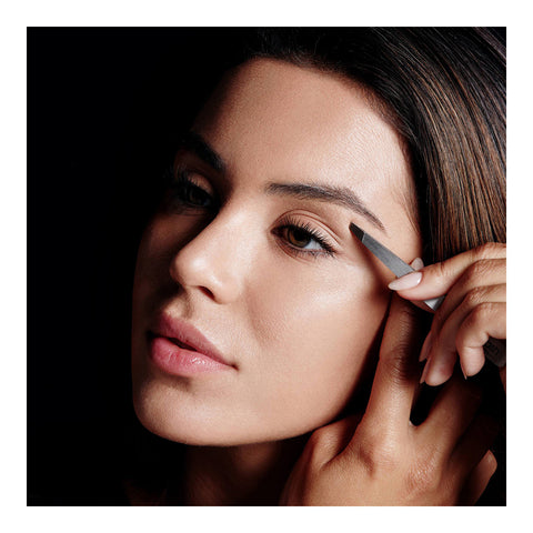 A woman with brown hair expertly shapes her eyebrows using the RevitaLash Precision Tweezer against a dark background, achieving groomed brows.