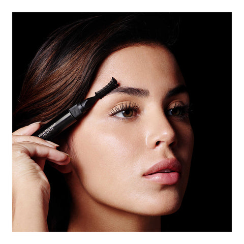 A woman with long dark hair uses the black RevitaLash Hi-Def Brow Gel to groom her eyebrows for precision styling. She has a neutral expression and smooth skin, set against a black background.